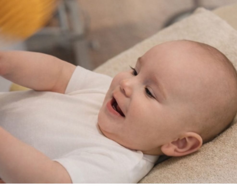 Ein lächelndes Baby in einem weißen T-Shirt liegt auf einer matten Unterlage.