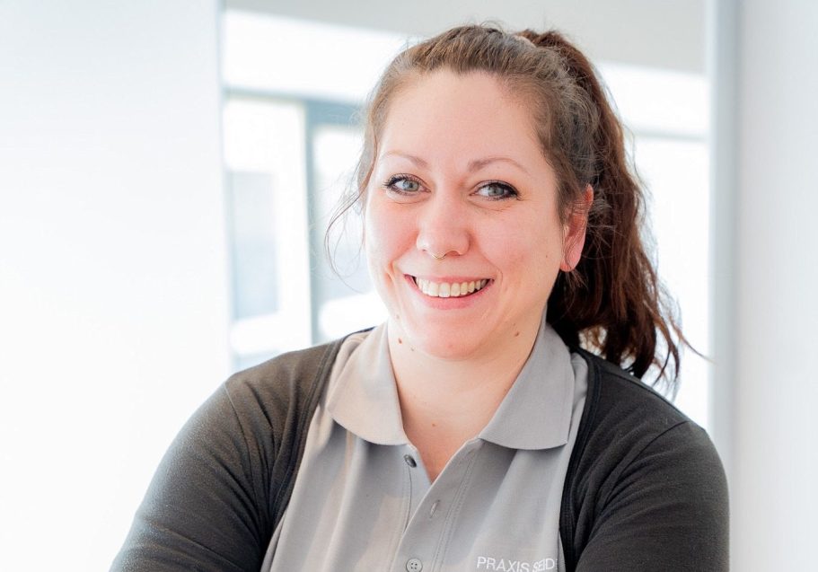 Frau mit langen, braunen Haaren, lächelt freundlich in einem hellen Raum.