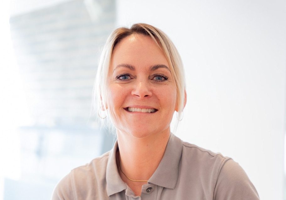 Lächelnde Frau mit blonden Haaren und grauem Oberteil in hellem Raum.