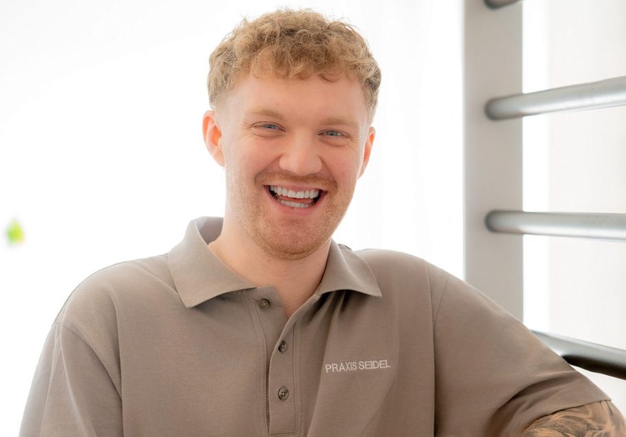 Lächelnder junger Mann mit blonder, lockiger Frisur, in einem grauen Polohemd.