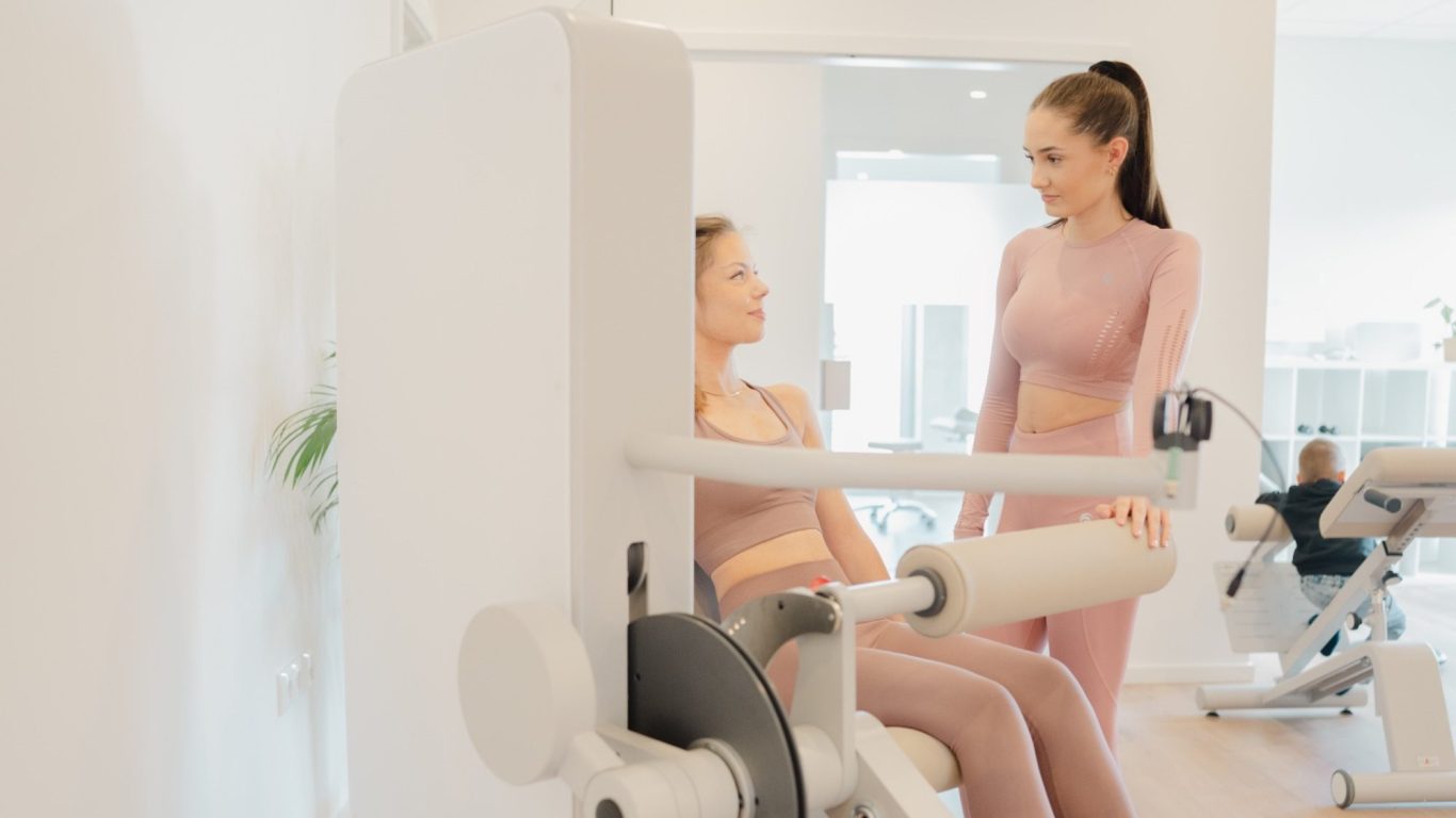 Zwei Frauen in Sportkleidung, eine berät die andere an einem Fitnessgerät.