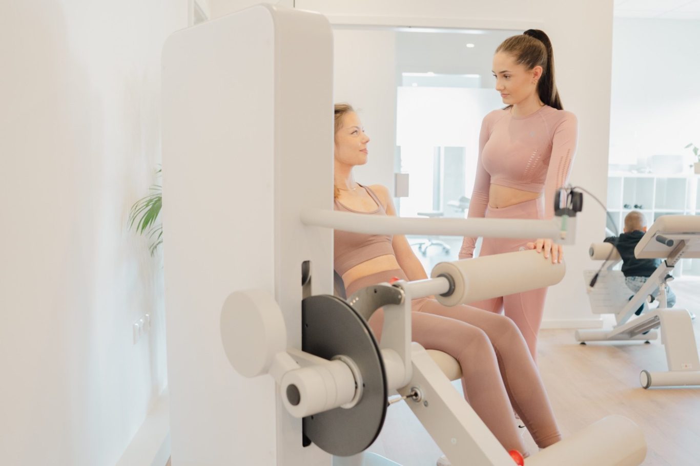 Zwei Frauen im Fitnessstudio, eine trainiert an einem Gerät, die andere gibt Tipps.