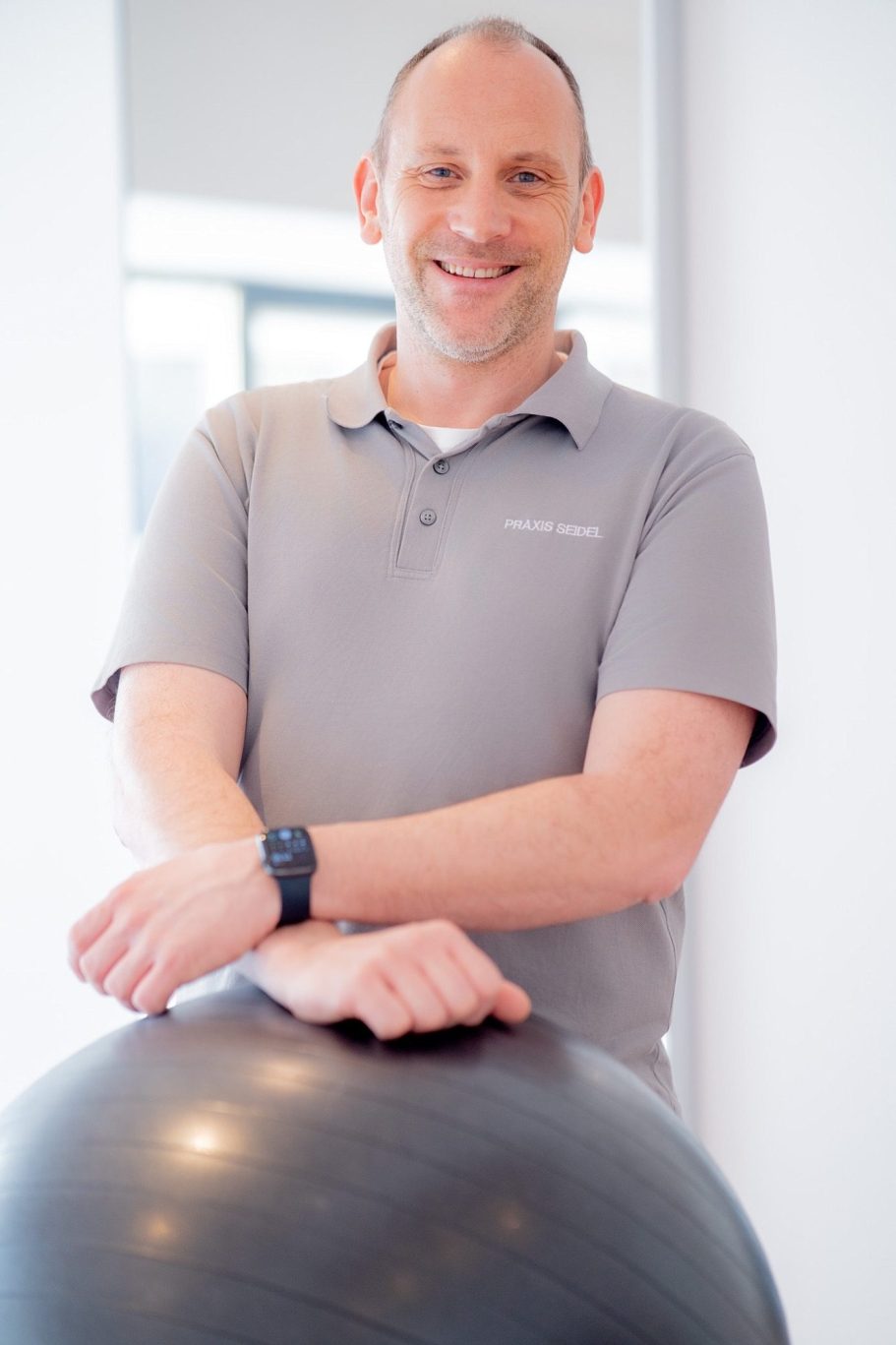 Mann lächelt, steht an einer Fitnessball und trägt ein graues Polohemd.