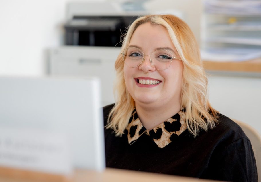 Lächelnde Frau mit blonden Haaren und Brille an einem Empfangstresen.