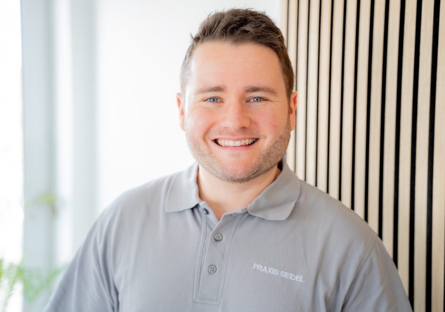 Lächelnder Mann in einem grauen Polohemd vor einer hellen, modernen Wand.