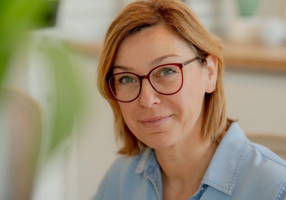Frau mit blonden Haaren und roten Brillen sitzt lächelnd am Tisch.