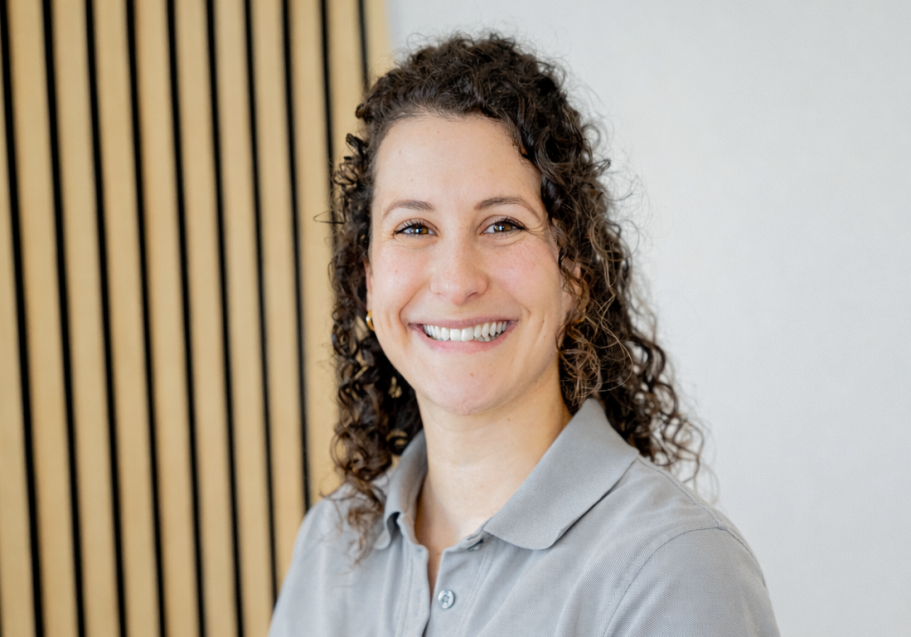 Lächelnde Frau mit lockigem Haar, hellgrauem Polo und neutralem Hintergrund.