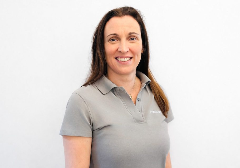 Frau mit langen, braunen Haaren trägt ein graues Polo-Shirt und lächelt freundlich.