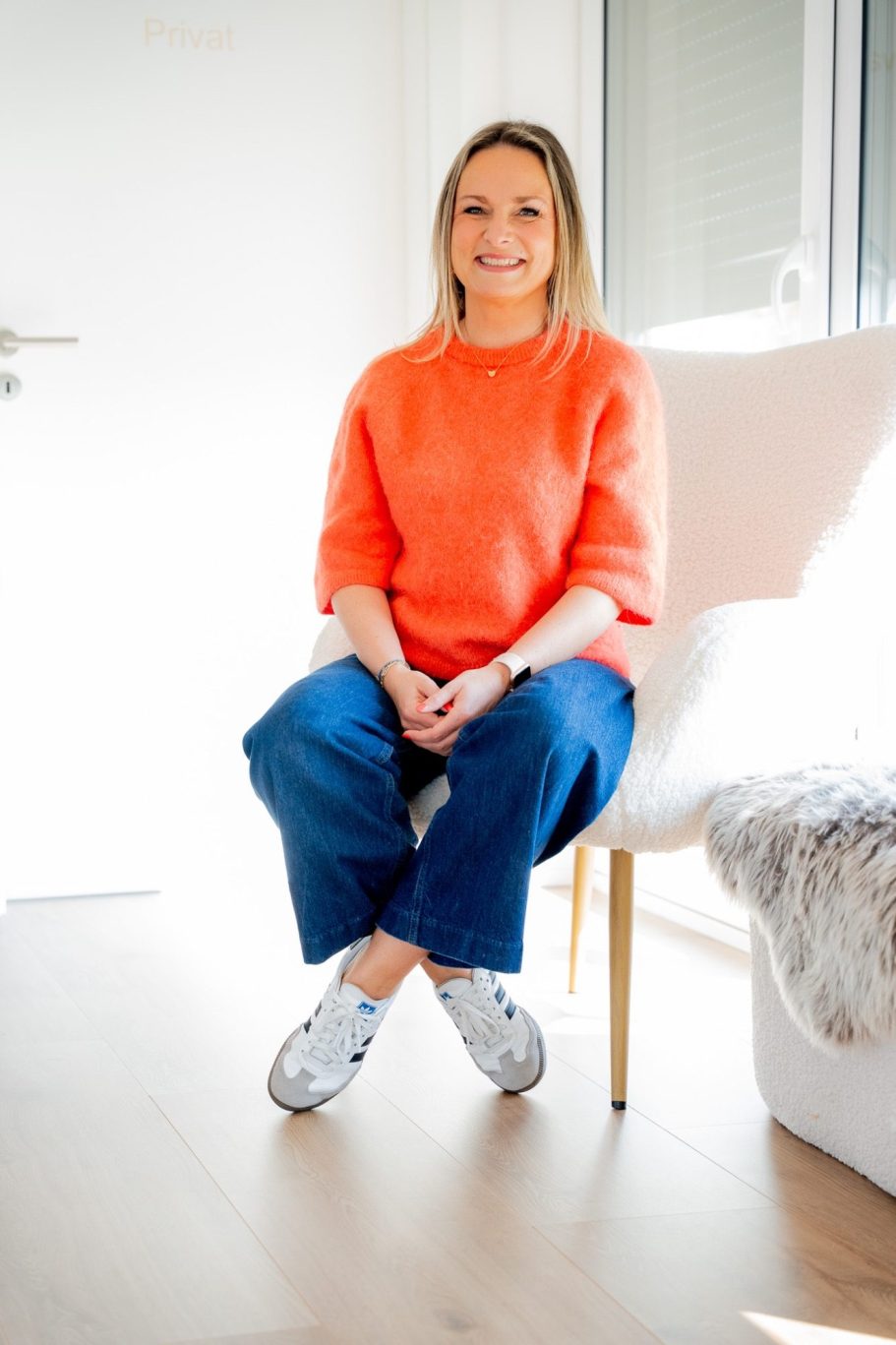 Frau mit blonden Haaren sitzt in einem hellen Raum auf einem Stuhl, trägt einen orangefarbenen Pullover.