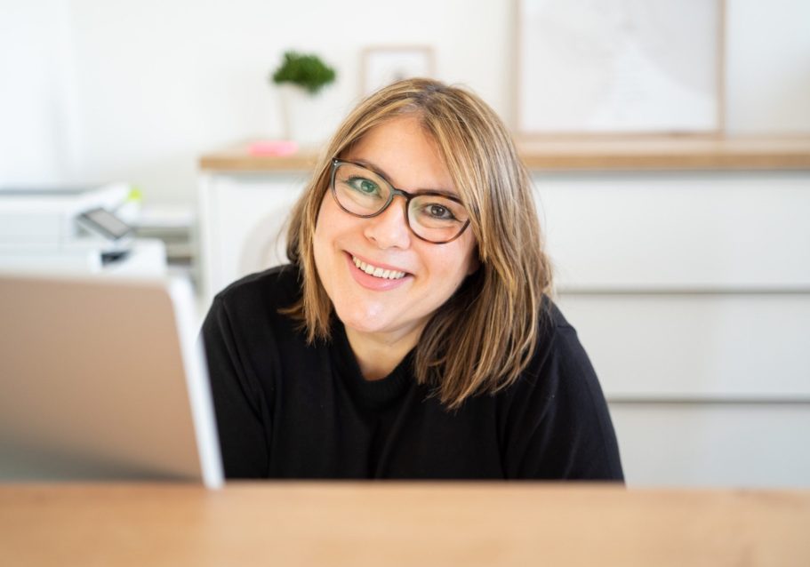 Lächelnde Frau mit Brille sitzt an einem Schreibtisch vor einem Laptop.