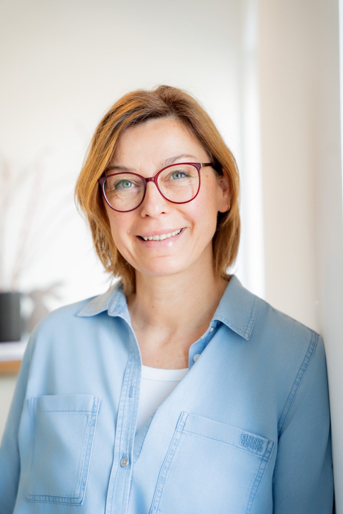 Frau mit blonden Haaren und Brille, die in einem hellen Raum lächelt.
